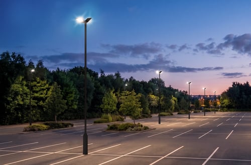 Empty commercial parking lot at dusk illuminated by pole-mounted area lights, providing even lighting across parking spaces and surrounding landscaping.